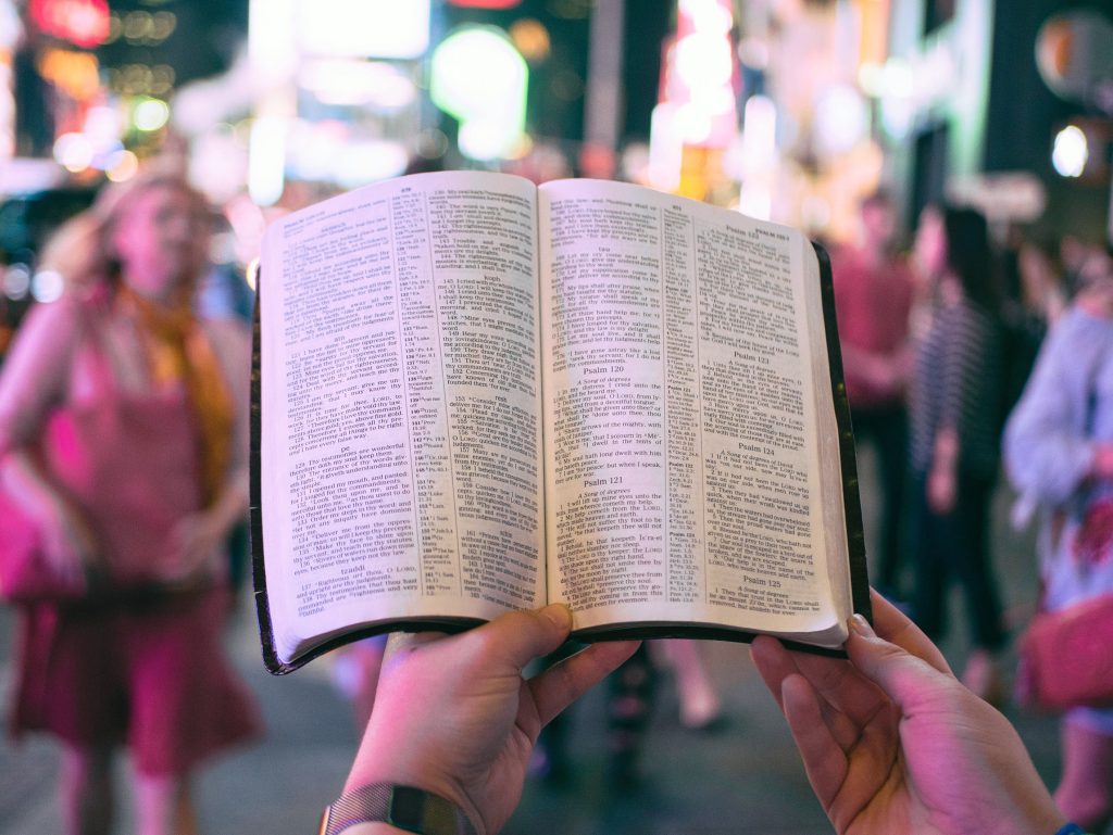 A photo of someone holding the Bible in the street with a view in the background of young people out on the town at night.